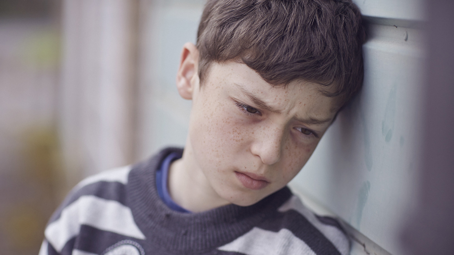 A young boy leans against a wall. His brow is knitted and he stares into the distance.