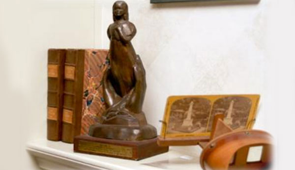 Wood sculpture of two large hands coming out of the ground, holding or molding the shape of a woman.