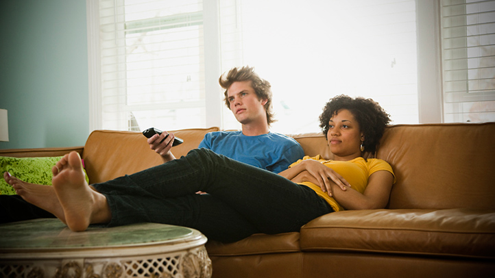 a man and woman sit on a sofa watching television.