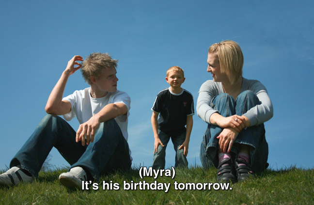 Three teens sitting on grass. caption: (Myra) It's his birthday tomorrow.