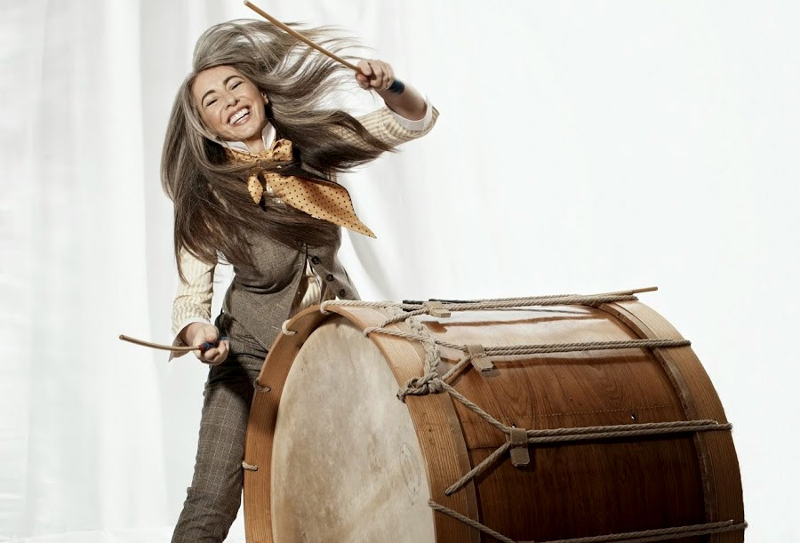 Evelyn Glennie joyously strikes a very large drum that is on it's side.