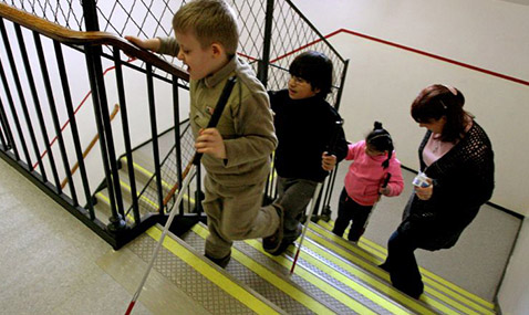 Children walk up stairs in a school stairwell. A boy with a white cane reaches the top.
