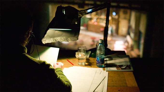 A woman sits at a desk and reads a printed script.
