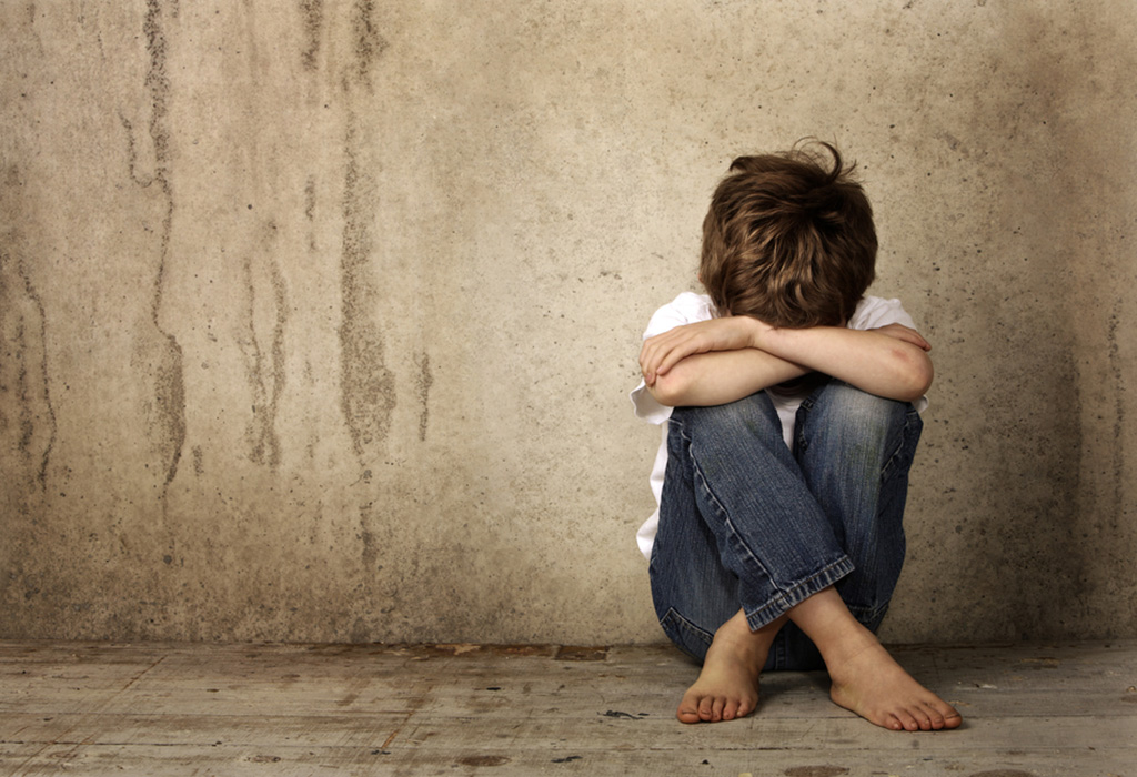 a child sits on the floor, arms on their raised knees, against a wall with their head down.
