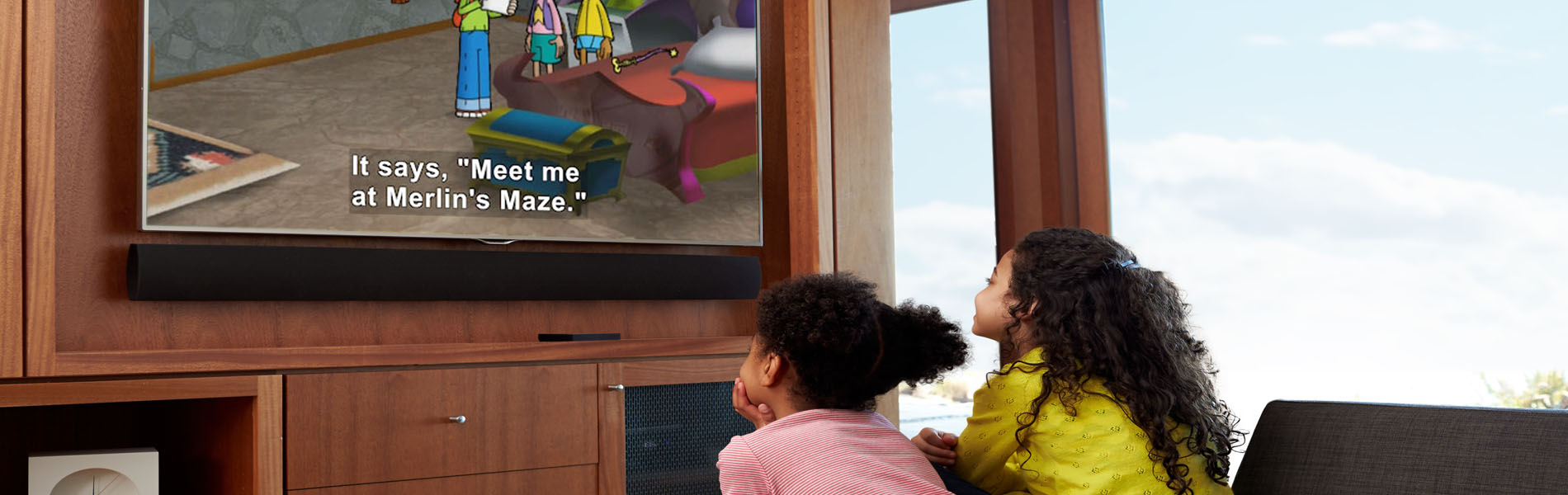 two young african american girls sit in front of a large tv on the wall,  watching an animated program with captions.