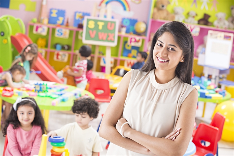 A smiling teacher stands in her classroom with young students behind her.