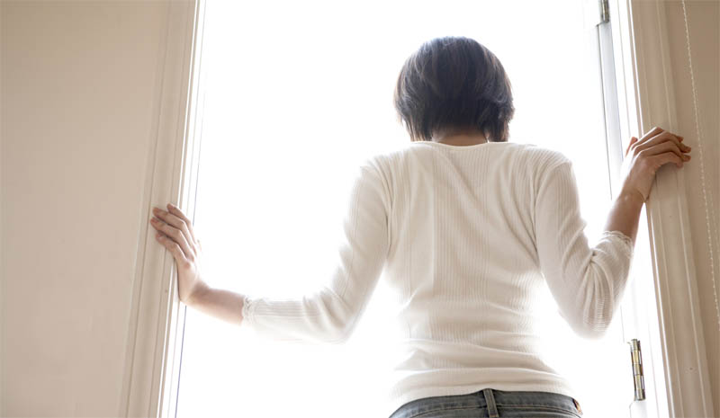 Back view of a young woman standing in a doorway with her hands on each side of the doorway. A bright light shines outside.
