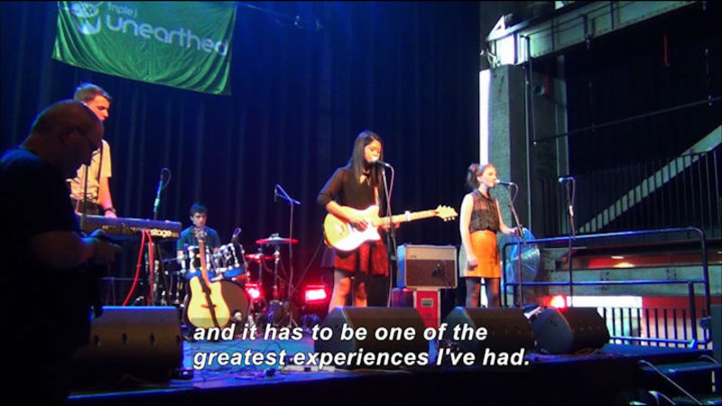 Screen capture of video. Four young people are on a stage playing musical instruments and singing. Caption reads 'and it has to be one of the greatest experiences I've had.'