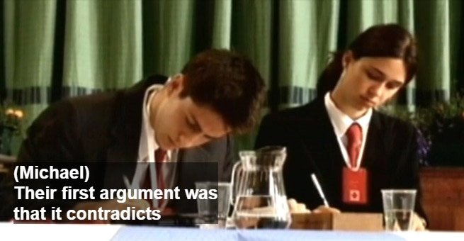Screen capture of video. Two debate students in suits sit at a table writing.  Captions read 'Michael: Their first argument was that it contradicts'