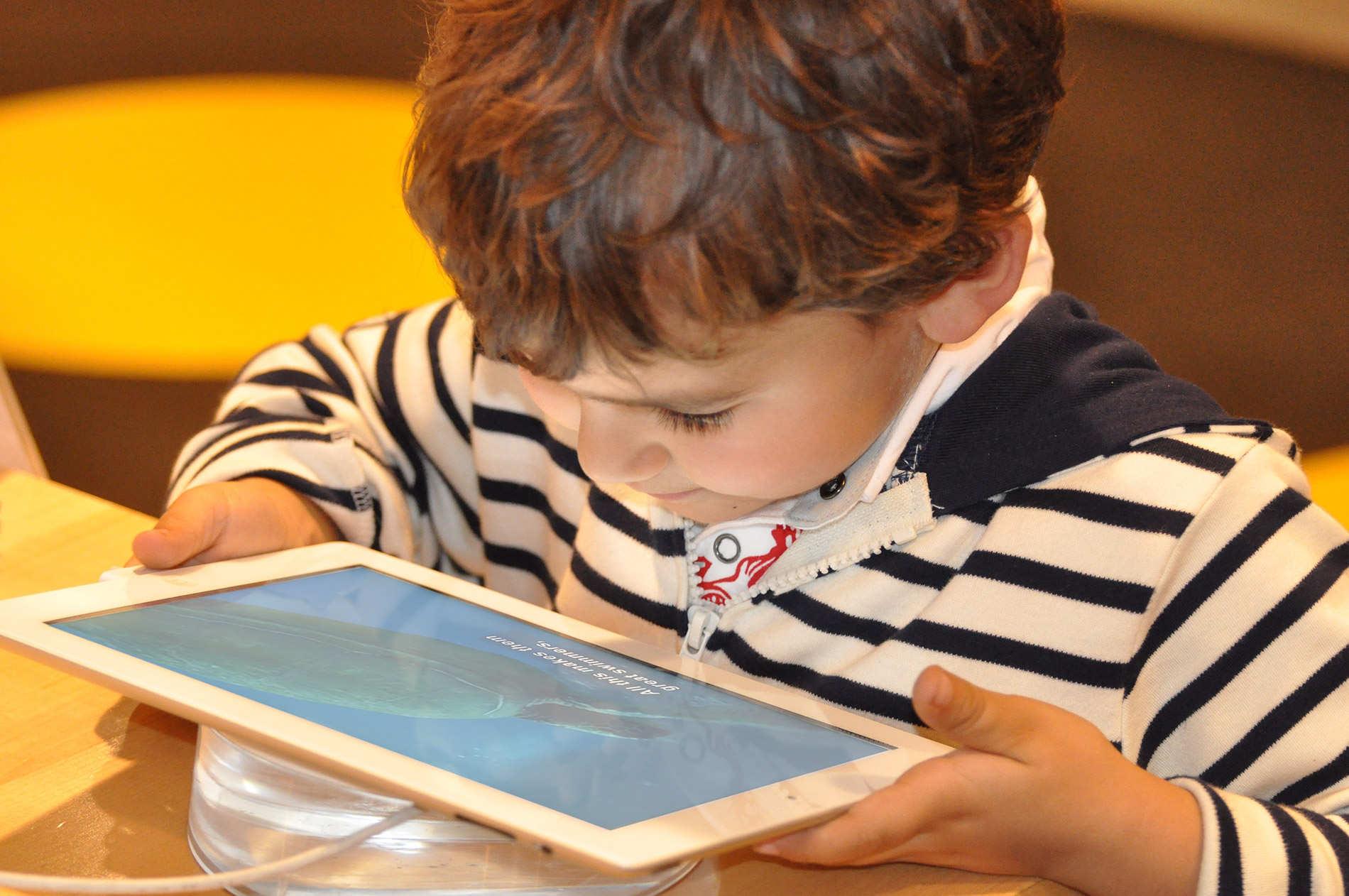 A young boy sits at a table, holding a tablet and watching a video on it.