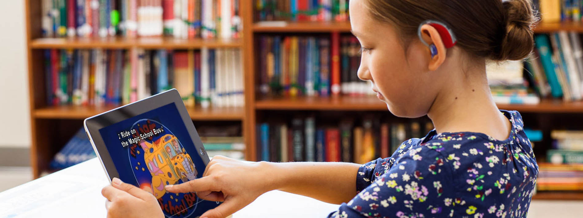 A young girl wearing a hearing aid sits at a tablet watching a captioned video on a tablet. Shelves of books are in the background.