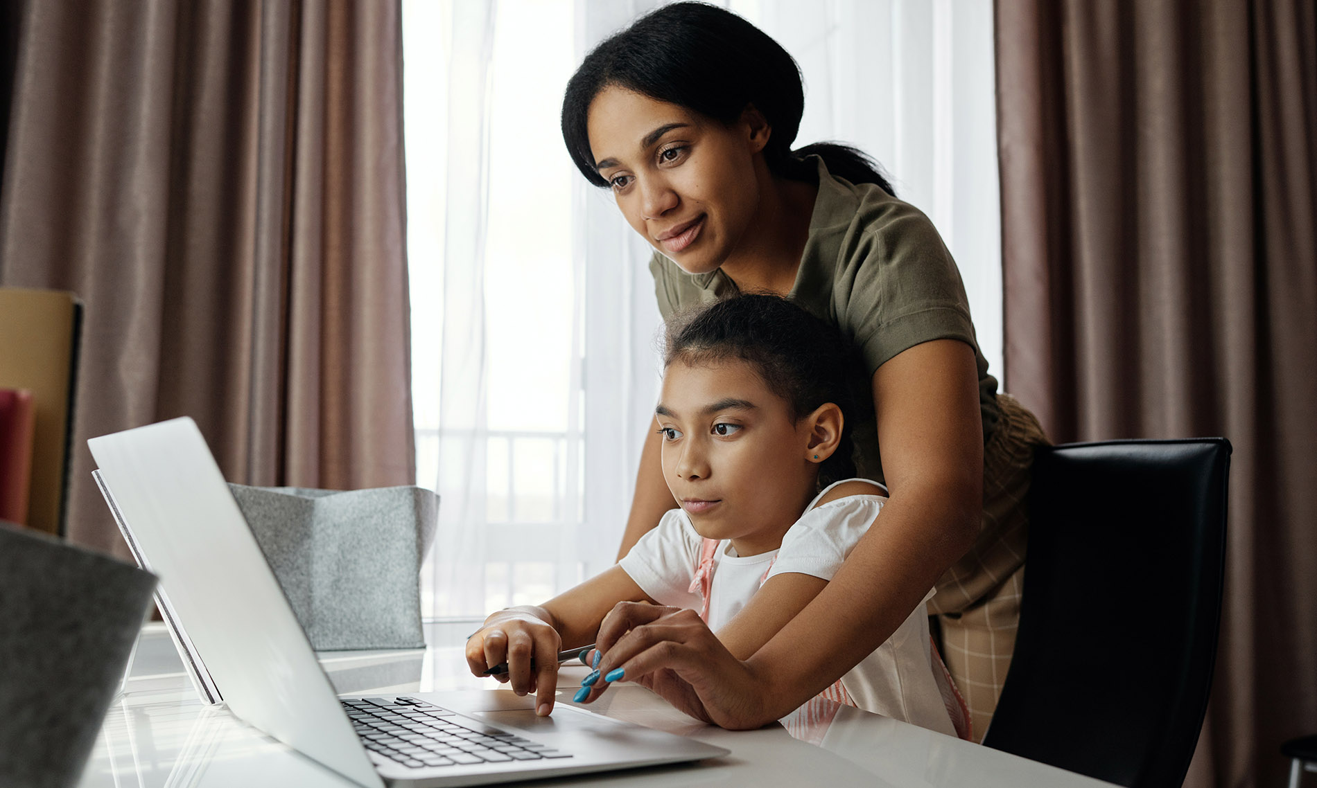 A mother stands behind her daughter who is seating at a desk at home. They are working on a laptop together.