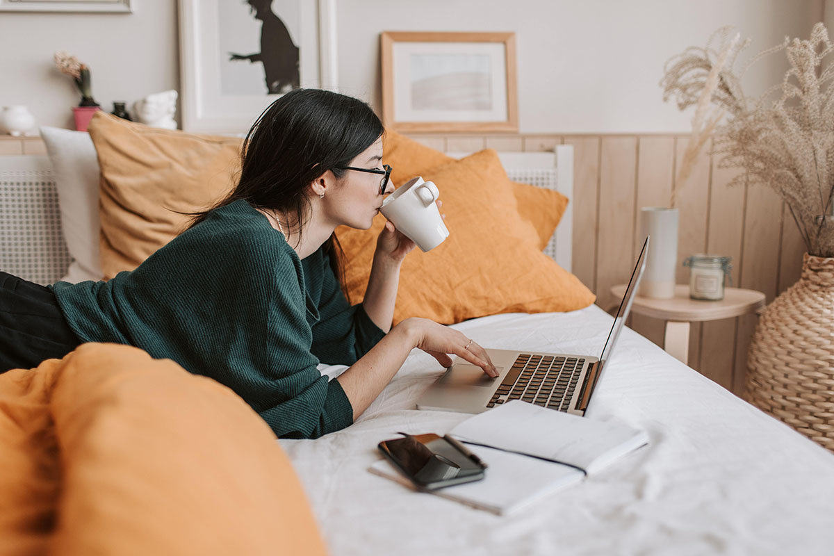 A woman lies on a bed while sipping from a coffee cup and working on a laptop.