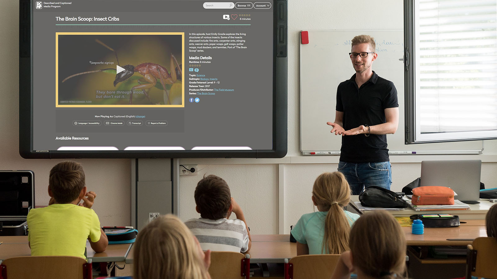 Teacher in a classroom with students, showing DCMP accessible video on a large screen.