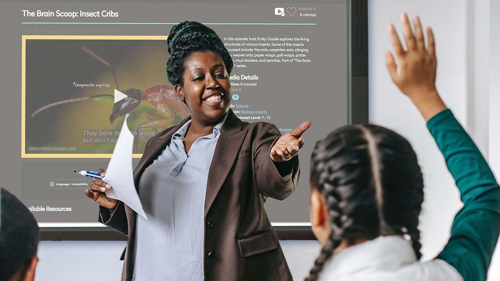 An African American teacher smiles and calls on one of her students who has her hand raised. A DCMP video is on the screen of a large smartboard behind her.