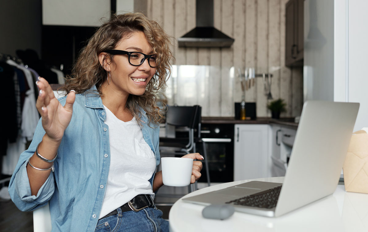A woman of color smiles and uses a laptop in her kitchen.