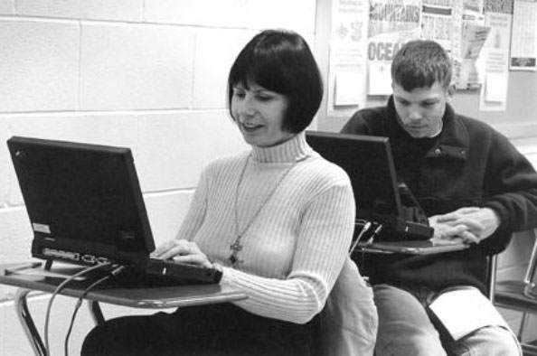 A woman and a man sit at desks in a classroom.