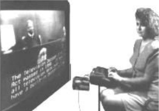 a woman sits at a desk typing as she watches a TV.