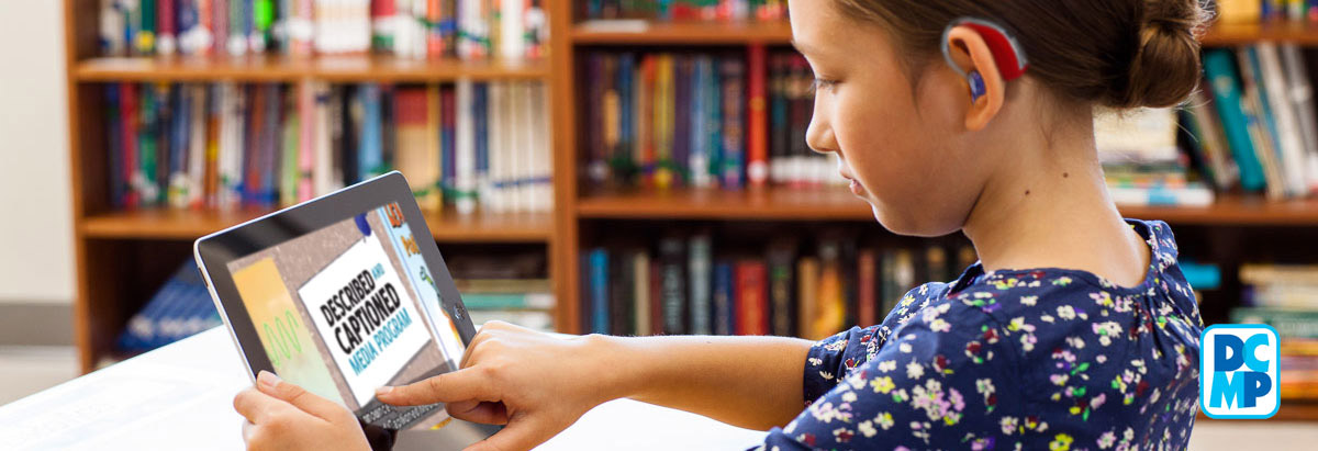 A young girl wearing a hearing aid sits in a school library watching DCMP media on a tablet computer.