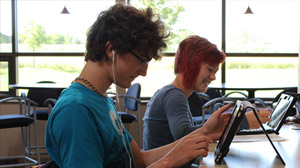 A teen boy and girl sit in a school library with computer tablets.