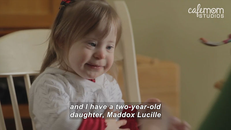 A young girl with Down Syndrome sits in a chair.  Caption reads and I have a two-year-old daught, Maddox Lucille