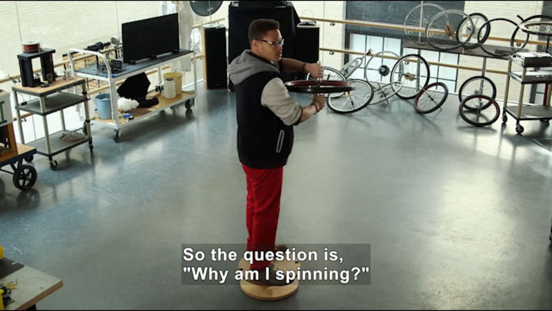 Person standing on a round platform holding a wheel. Caption: so the question is, "Why am I spinning?"