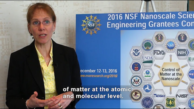 Person speaking and standing next to a poster board with "Control of matter at the nanoscale" surrounded by logos from different organizations. Caption: of matter at the atomic and molecular level.