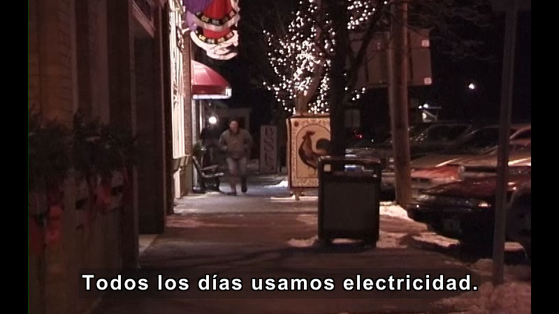 City sidewalk with well-lit buildings lining one side. Spanish captions.