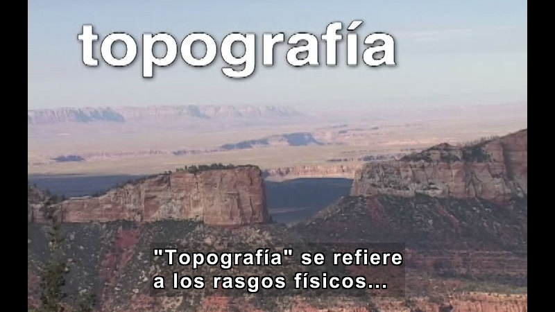 Rock formations with straight sides rise above rocky red landscape dotted with trees. Spanish captions.