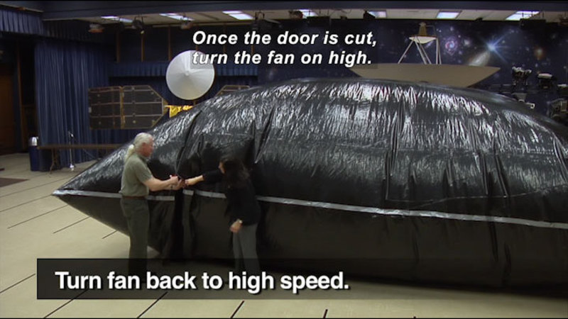 People standing in front of a giant inflated black plastic pouch. Turn fan back to high speed. Caption: Once the door is cut, turn the fan on high.