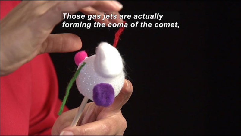 Person attaching different colored and shaped objects to a Styrofoam ball. Caption: Those gas jets are actually forming the coma of the comet,