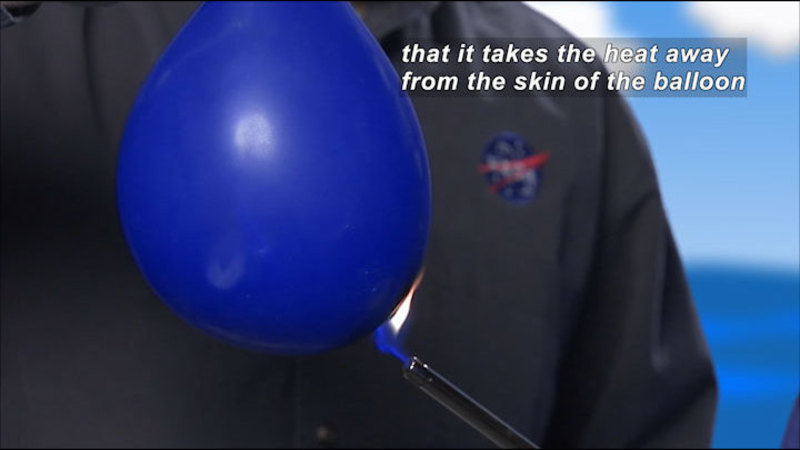 Person wearing a jumpsuit with a NASA logo holds a flame to an inflated balloon. Caption: that it takes the heat away from the skin of the balloon