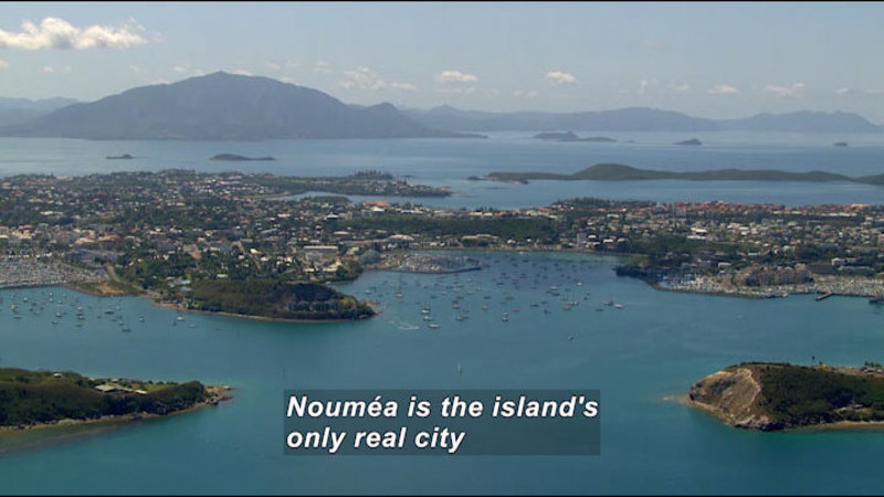 Aerial view of a populated island surrounded by bluish green water. Caption: Nouméa is the island's only real city