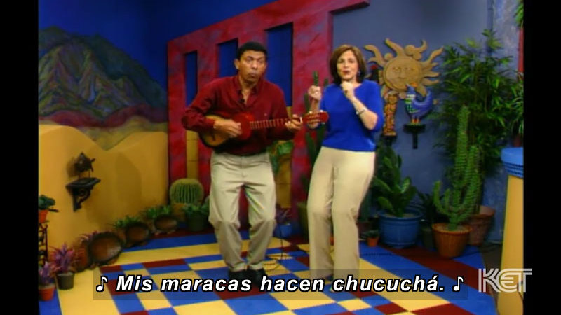 Two people in a colorful room with cacti in pots all around the walls. Spanish captions. One is playing an instrument.