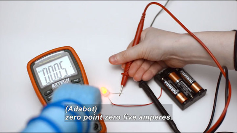 Measurement device placed on a circuit powered by three AA batteries. Device shows 0.05 on the display. Caption: (Adabot) zero point zero five amperes.