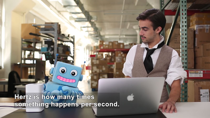 Person and a robot in a workshop with a laptop in front of them. Caption: Hertz is how many times something happens per second.