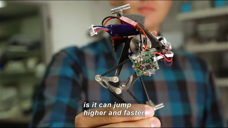 Person holding a small mechanical device with leg-like structures and a circuit board. Caption: is it can jump higher and faster