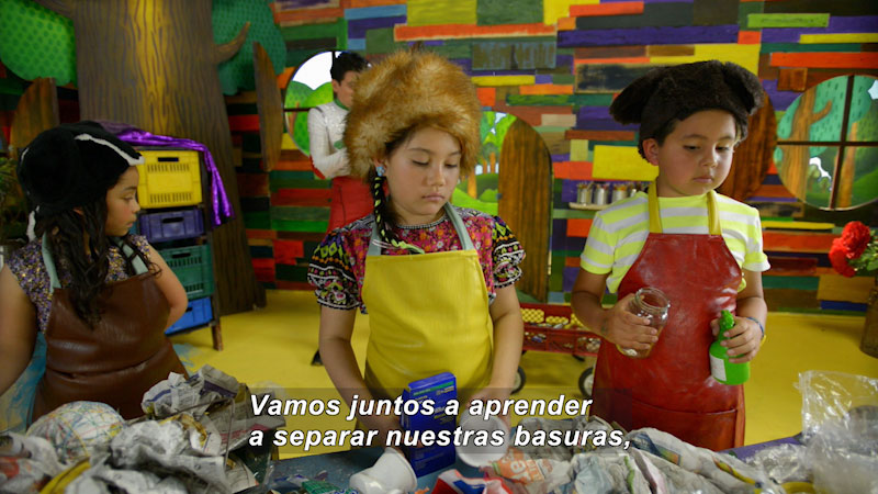Children wearing aprons standing in front of table covered in trash. Spanish captions.