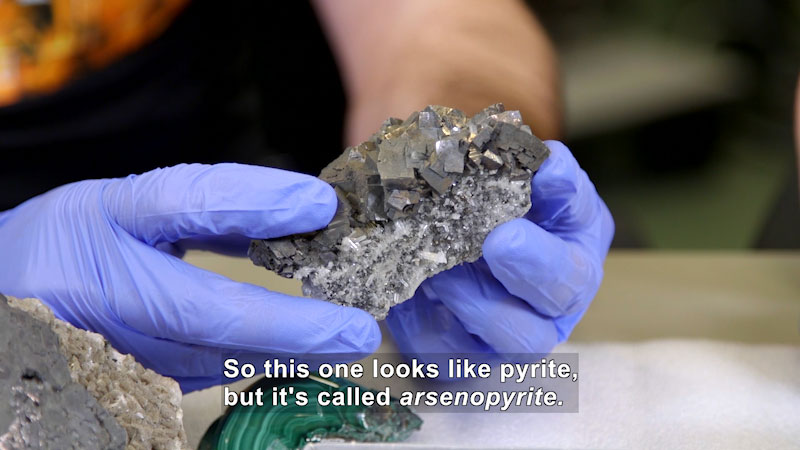 Person holding a silvery rock with geometric chunks on top and white and silver crystalline structures on the bottom. Caption: So this one looks like pyrite, but it's called arsenopyrite.