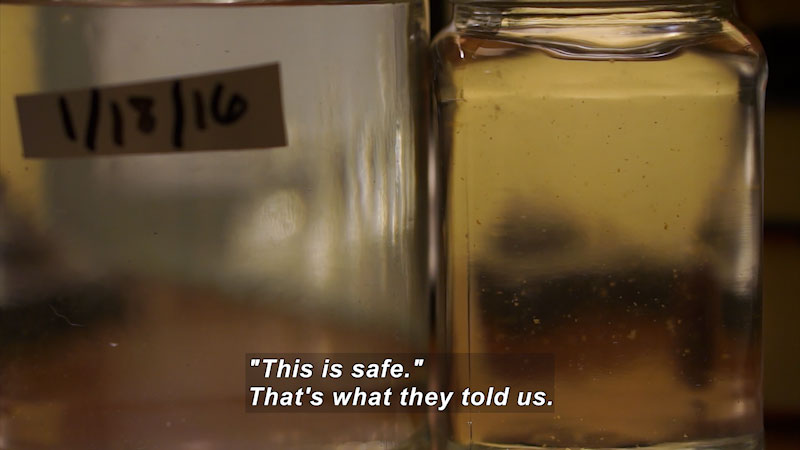 Closeup of glass containers of water with visible particulate matter. One is labeled 1/18/16. Caption: "This is safe." That's what they told us.