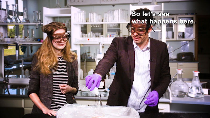 Two people in a lab. One is pouring liquid into a beaker. Caption: So let's see what happens here.