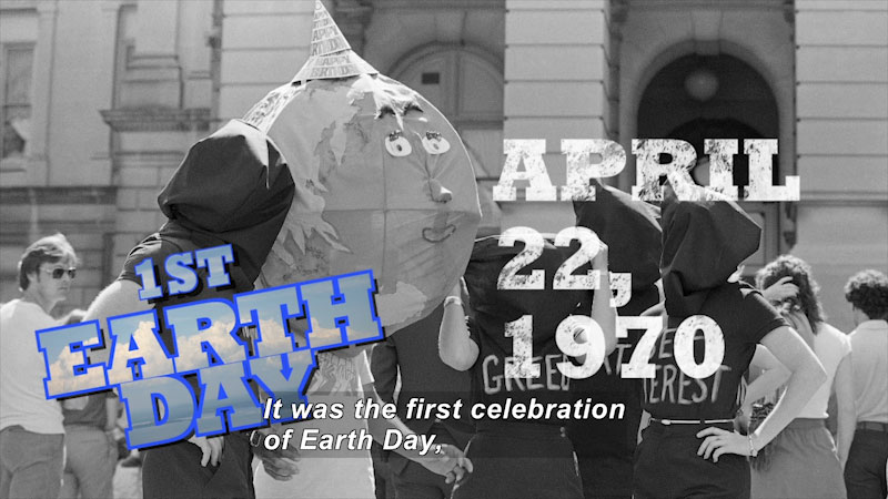 People with sacks covering their faces and phrases such as "Greed" and "Self Interest" written on their shirts stand next to a person wearing Earth with a face over their head. 1st Earth Day April 22, 1970 Caption: It was the first celebration of Earth Day,
