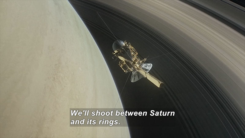 A cylindrical space craft with equipment and antennas protruding from it in relief against a large grayish-brown planet with rings in the background. Caption: We'll shoot between Saturn and its rings.