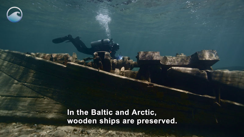 Scuba diver swimming above a wooden ship submerged in shallow water. Caption: In the Baltic and Arctic, wooden ships are preserved.