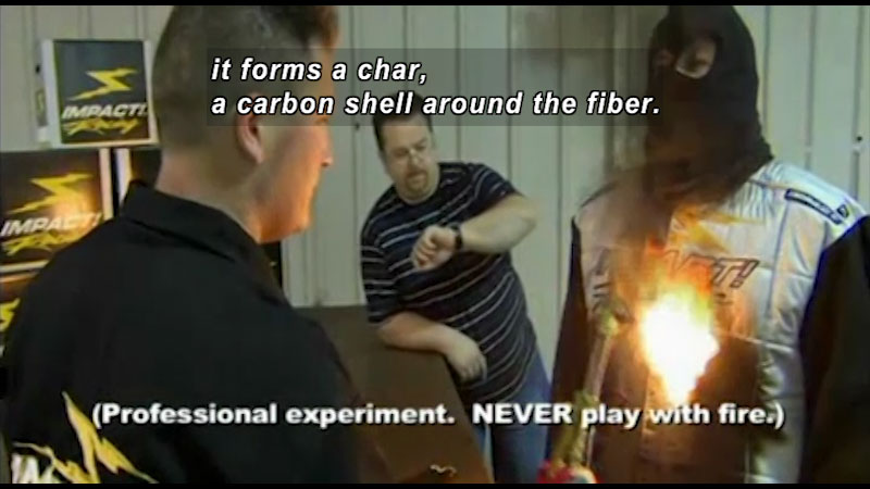 Three men, one wearing a vest being lit on fire. Caption: it forms a char a carbon shell around the fiber. 