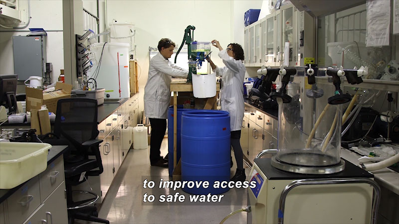 Two people in a science lab working on a piece of equipment from opposite sides of the table. The equipment terminates in a spout leading to a large plastic drum. Caption: to improve access to safe water