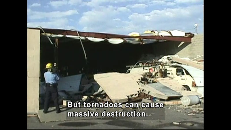 Partially collapsed building covering a vehicle in debris. A person wearing a hard hat films the destruction. Caption: But tornadoes can cause massive destruction.
