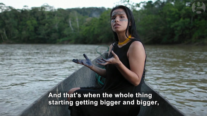 Indigenous person sitting in the bow of a canoe on the water with the jungle in the background. Caption: And that's when the whole thing started getting bigger and bigger,