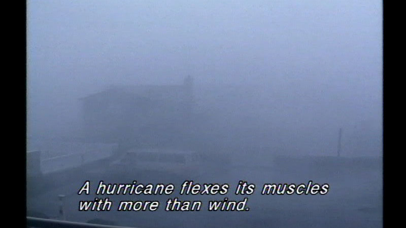 Buildings and a vehicle barely visible through a storm. Caption: A hurricane flexes its muscles with more than wind.
