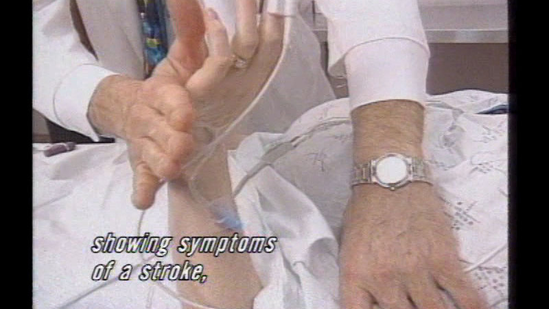 Torso and arm of a person with an oximeter and an IV in a hospital bed while a doctor tests responsiveness. Caption: showing symptoms of a stroke,